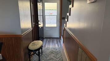 Un pasillo con una puerta que da a una habitación luminosa, un taburete pequeño y un termostato fijado a la pared.