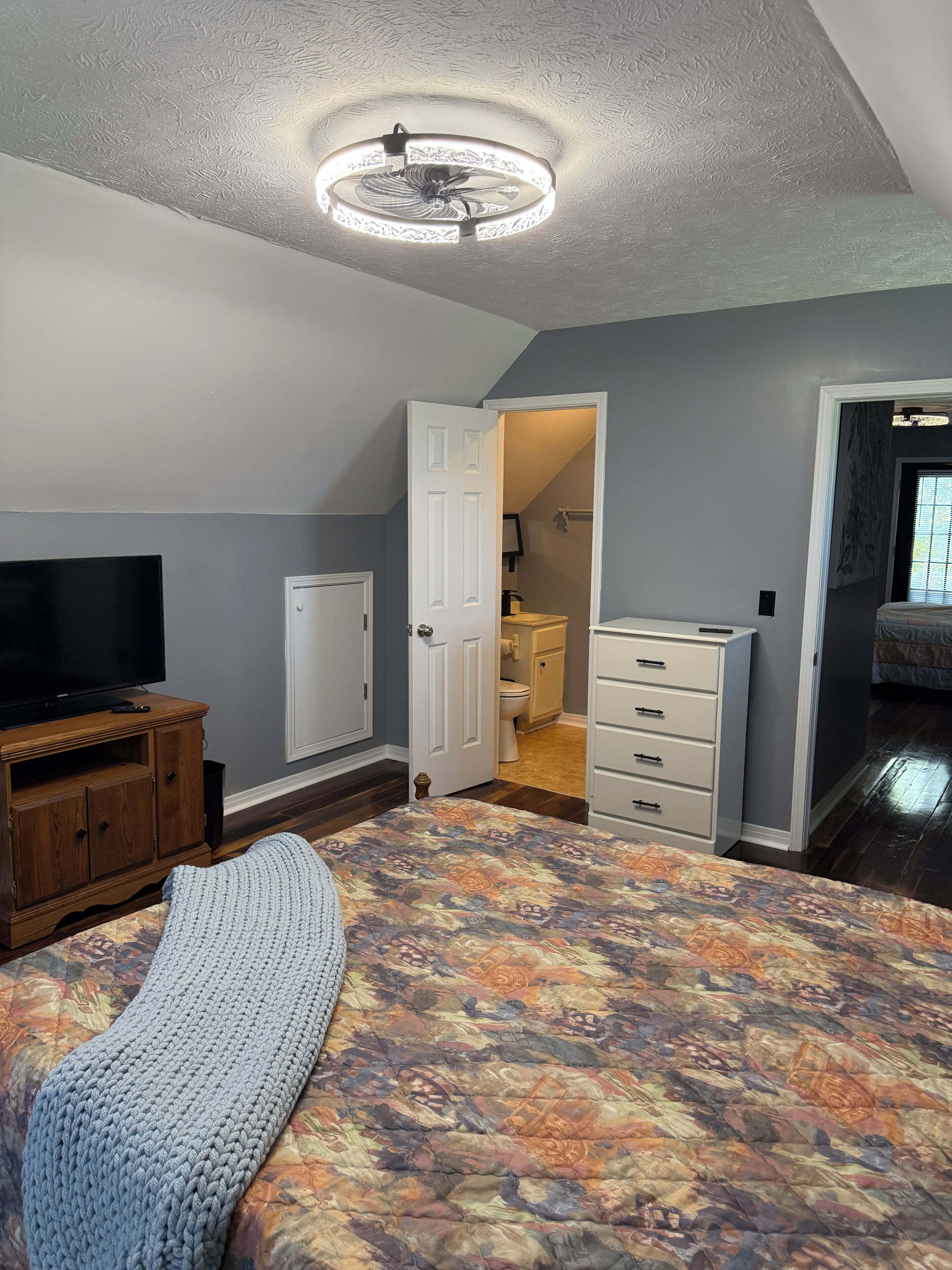 Un dormitorio con cama, televisor, cómoda y ventilador de techo.