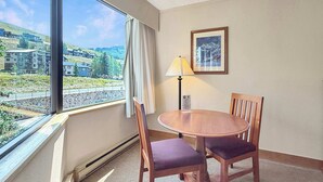 Hotellihuone, jossa on suuri ikkuna, pyöreä puupöytä ja kaksi tuolia, lamppu sekä seinällä oleva kehystetty kuva.