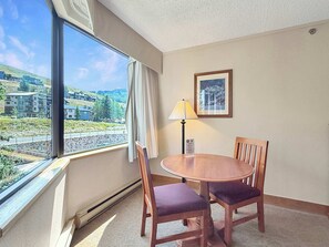 Quarto de hotel com uma grande janela, uma mesa redonda de madeira com duas cadeiras, um abajur e um quadro na parede.