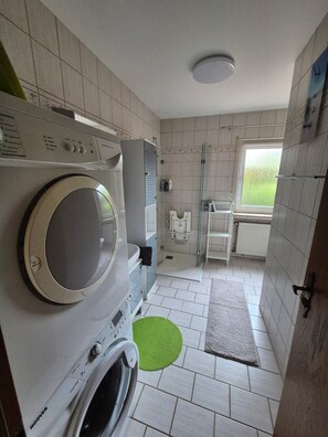 Een praktische badkamer met een wasmachine, een droger, een douchecabine van glas en een raam voor natuurlijk licht.