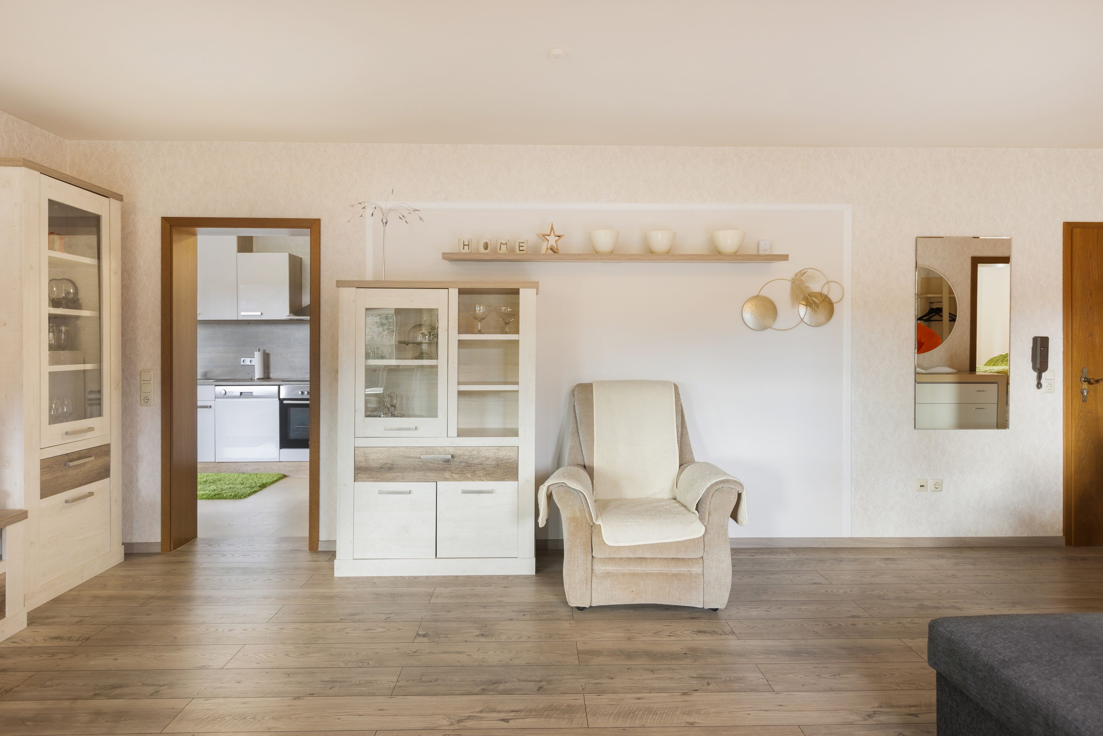 Een woonkamer met een beige fauteuil, een wit kastje met glazen deuren, en een keuken zichtbaar door een open doorgang.
