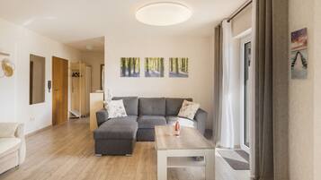Ein modernes Wohnzimmer mit einem grauen Ecksofa, einem Holztisch und einem großen Fenster mit Vorhängen.