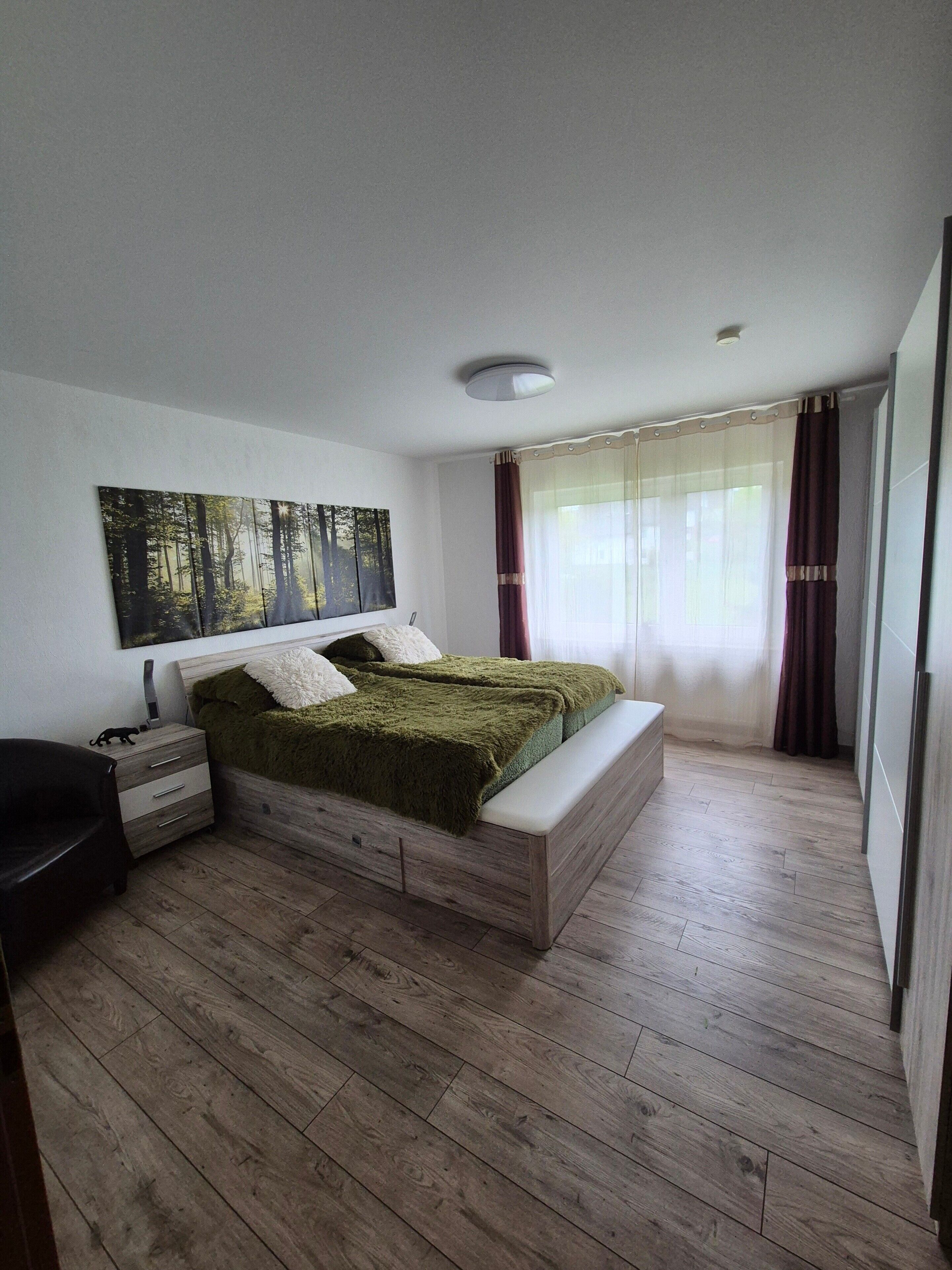 Een slaapkamer met een bed, nachtkastjes, een bankje, een wanddecoratie en een raam met gordijnen.