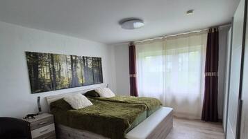 Een slaapkamer met een bed, nachtkastjes, een bankje, een wanddecoratie en een raam met gordijnen.