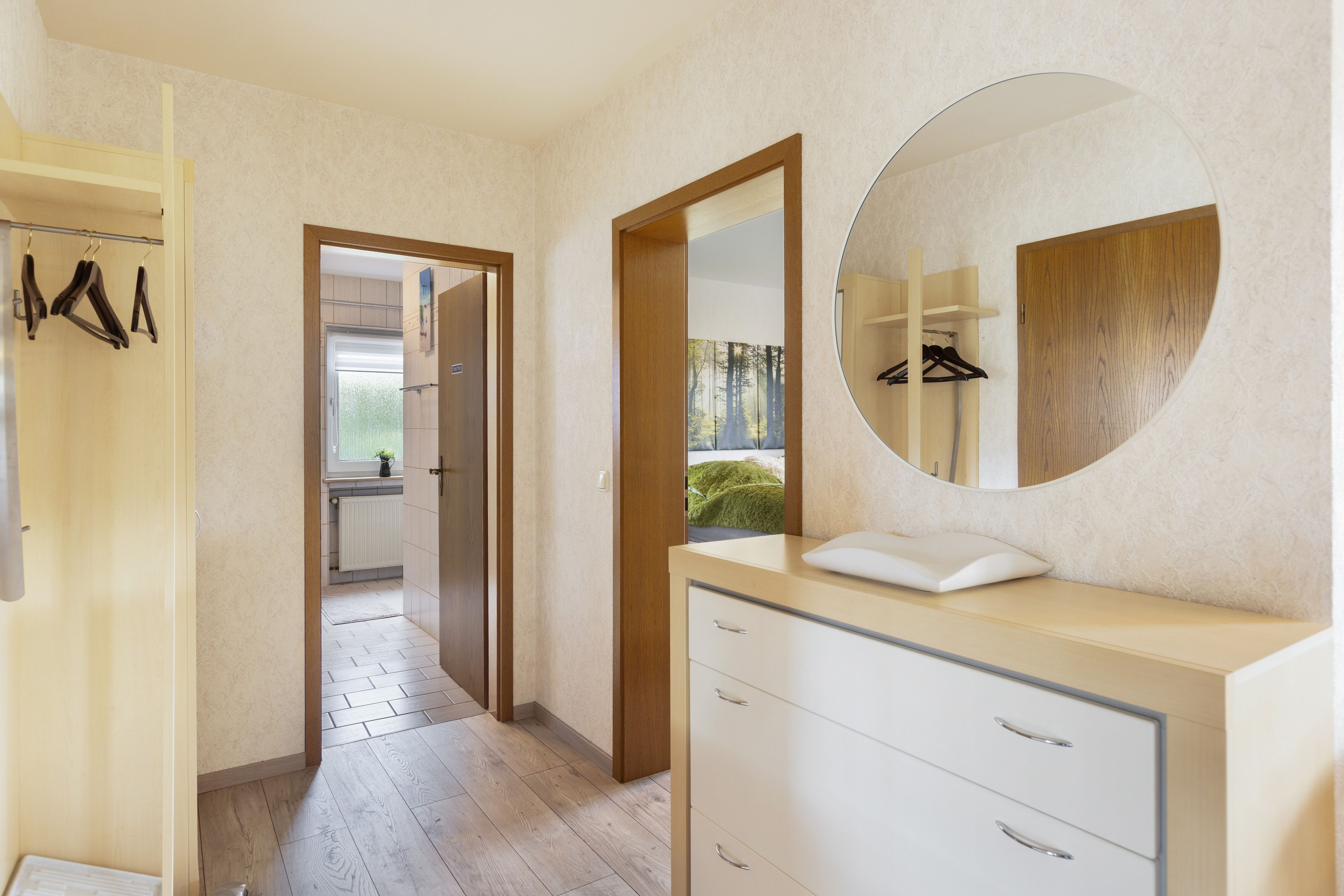 A modern bathroom with a wooden vanity, a round mirror, and a walk-in closet with hanging clothes.