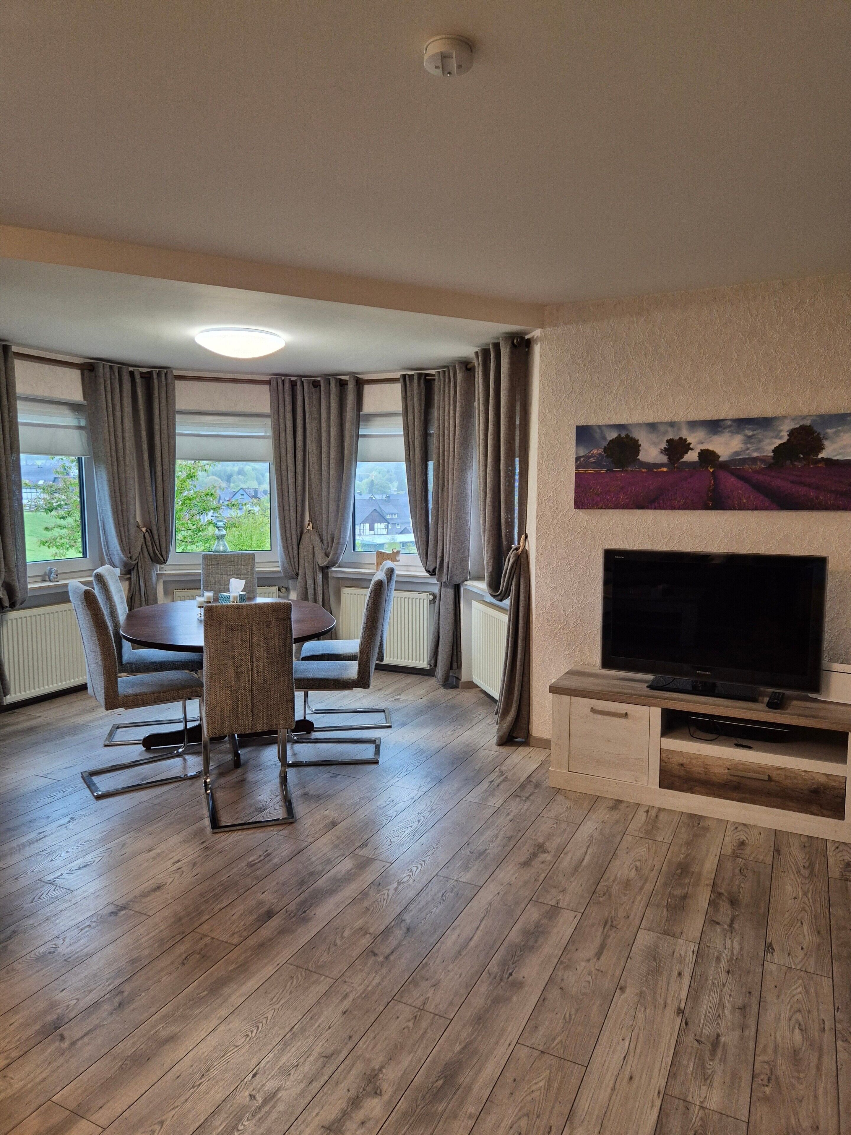 Een kamer met een eettafel, stoelen, een flatscreen televisie en een schilderij aan de muur.