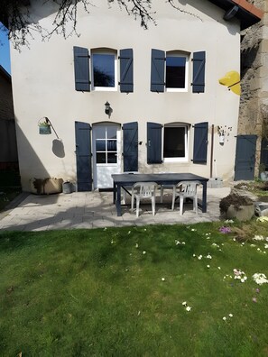Un bâtiment à deux étages doté d’un patio comprenant une table et des chaises, un petit jardin fleuri et un mur en pierre.