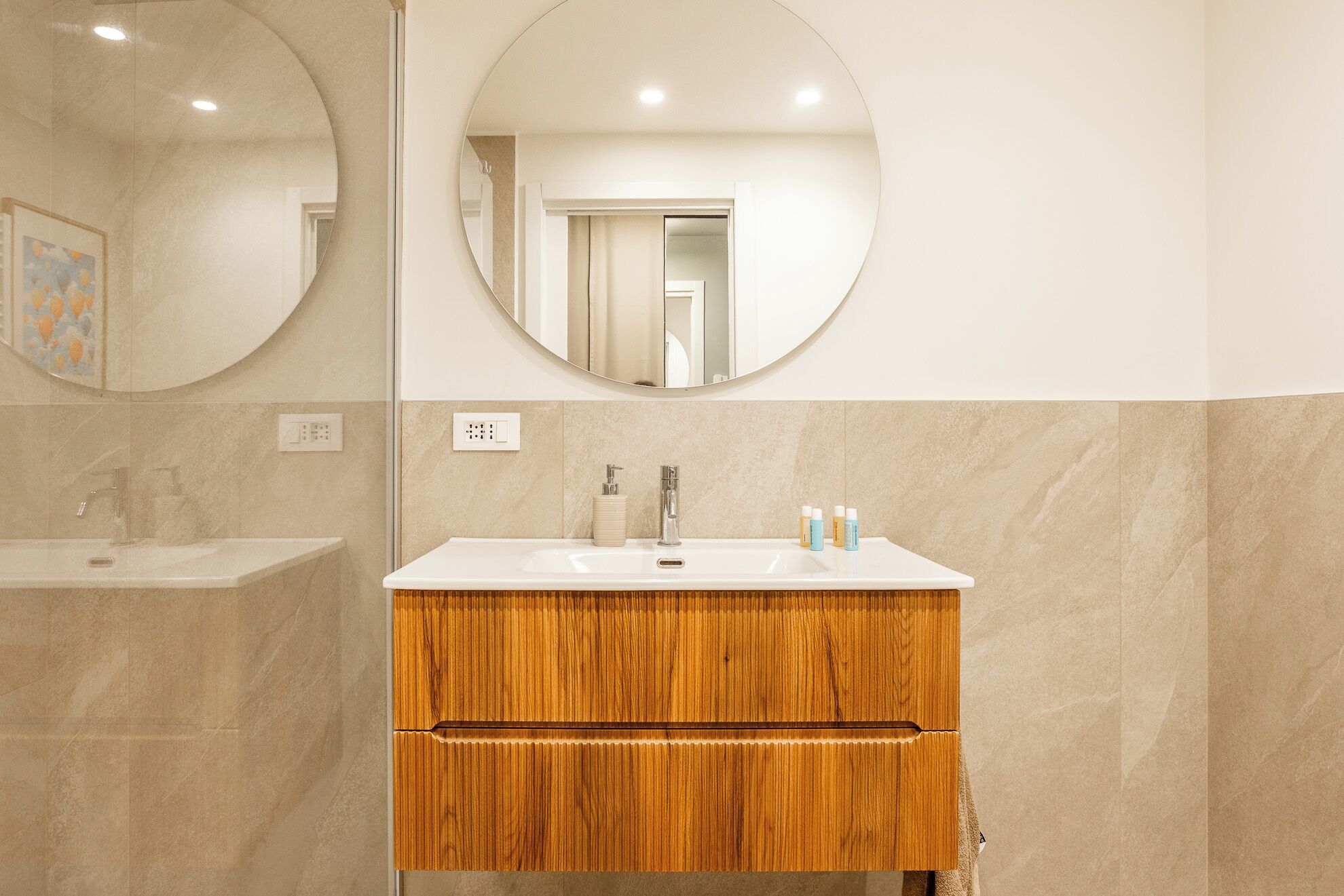 Un baño con lavamanos blanco, tocador de madera y espejo redondo.