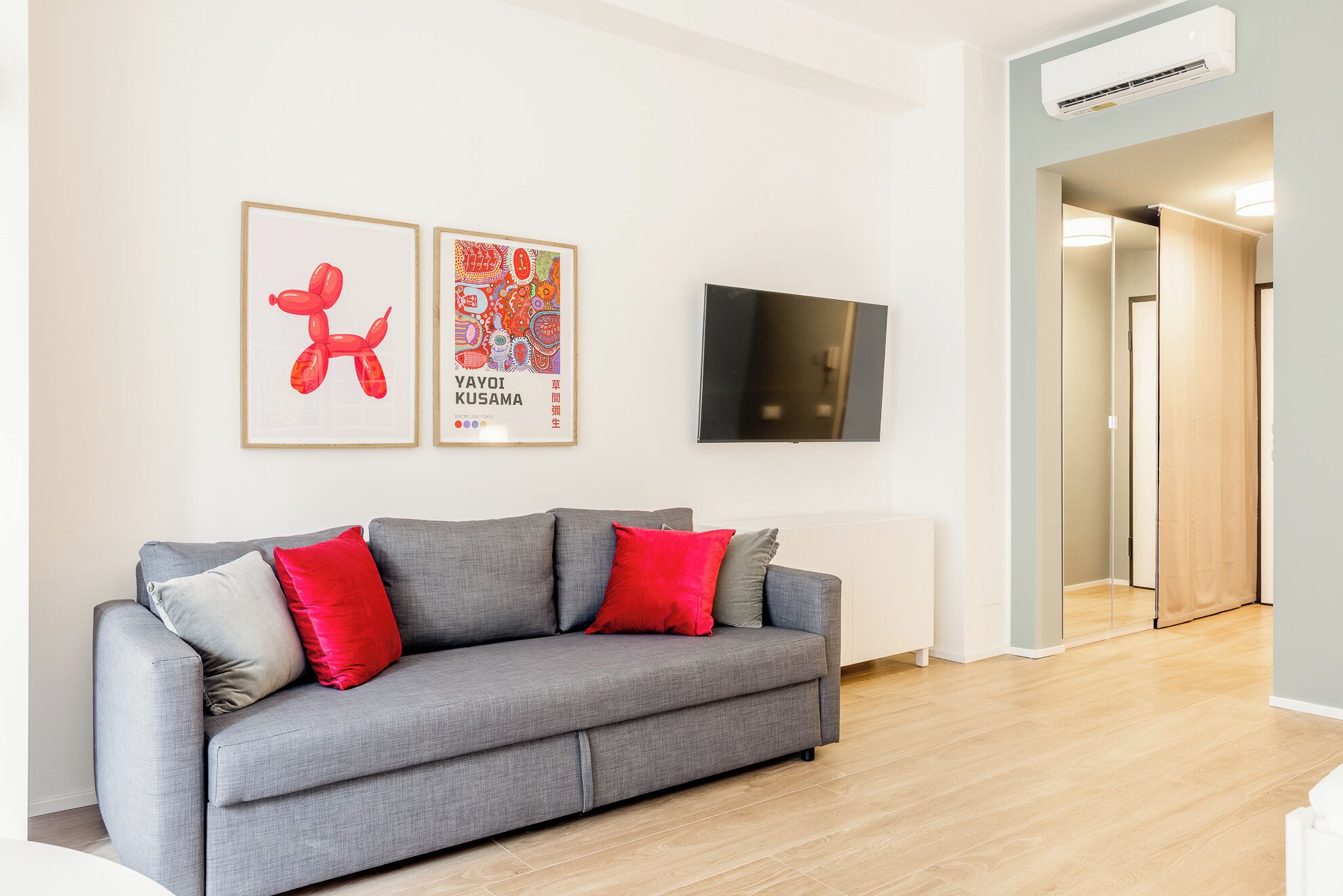 Een moderne woonkamer met een grijze bank, rode en grijze kussens, een flatscreen televisie en twee ingelijste kunstwerken aan de muur.