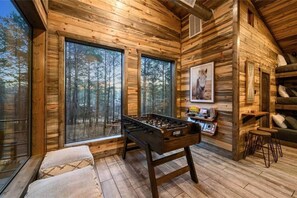 A wooden cabin interior with a foosball table, large windows, and a framed picture on the wall. - IIII Pines + a Cone: Sleeps 18, Hot Tub, Game Room (Hochatown)