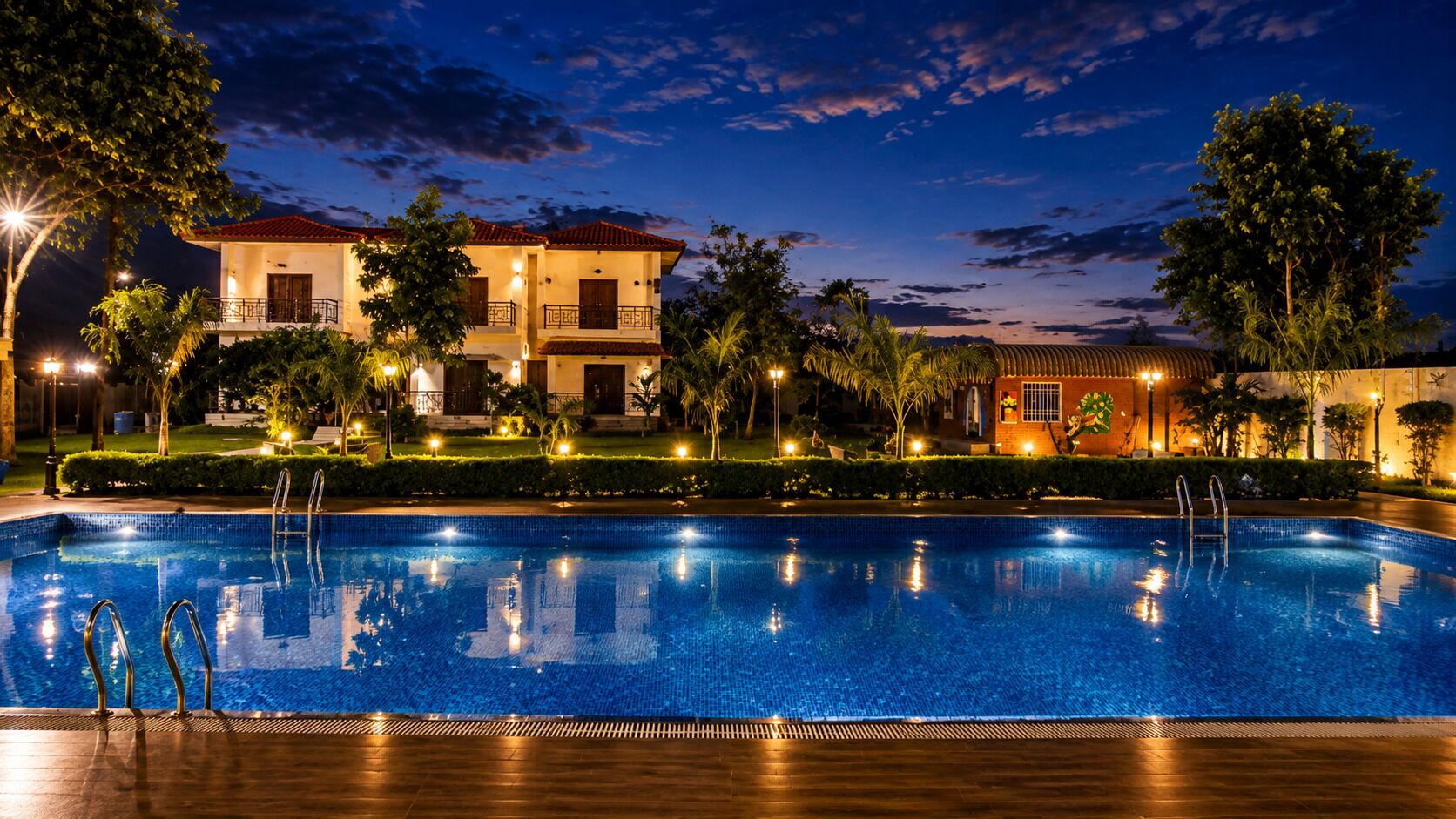 A swimming pool with a wooden deck, surrounded by well-lit buildings and palm trees.
