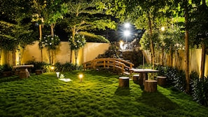 A well-lit garden with a wooden bridge, stone steps, and a small pond.
