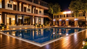 A swimming pool with a wooden deck, surrounded by a building with multiple floors and balconies. - Narayana Green By Clickon Resort (Sherghati)