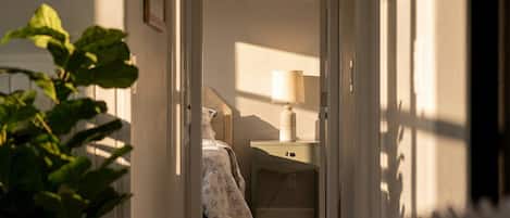A hallway leading to a bedroom with a bed, a bedside table with a lamp, and a wall-mounted shelf.