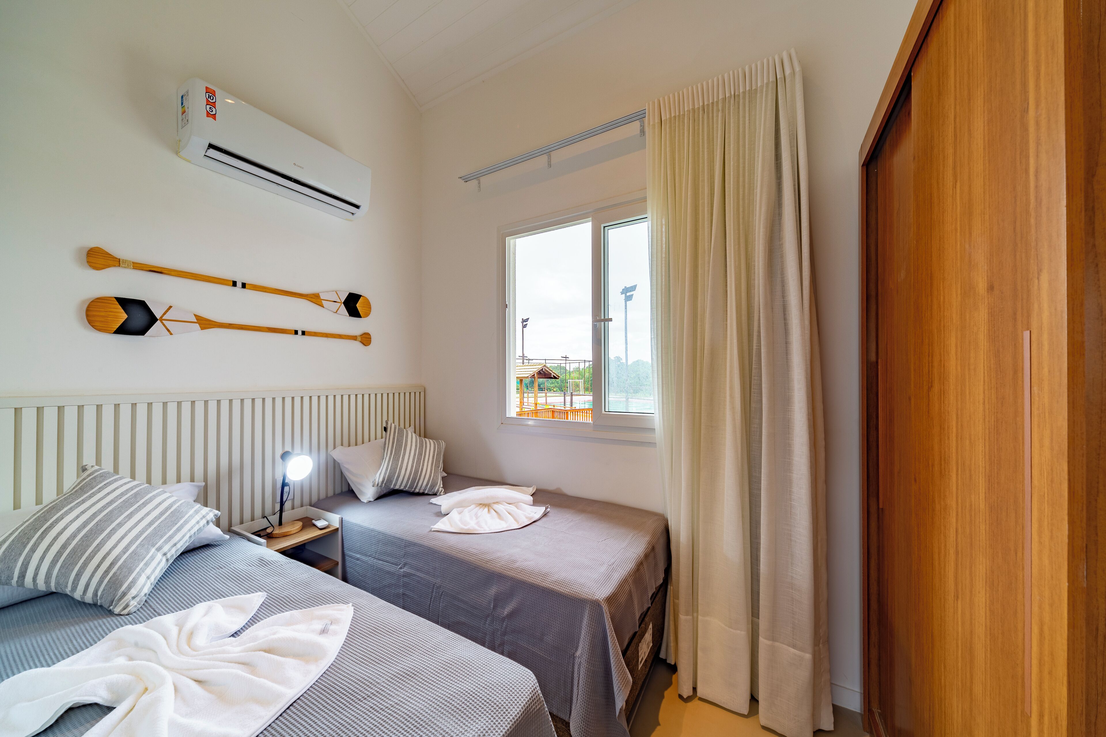 A hotel room with two beds, a window with curtains, a bedside table with a lamp, and a wall-mounted air conditioner.