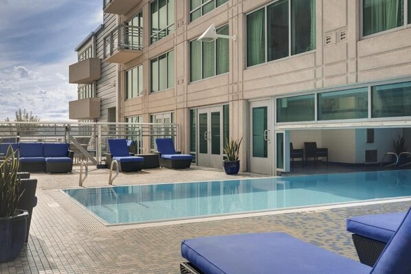Una piscina en la azotea con sillones de descanso y un edificio con cerramiento de vidrio.