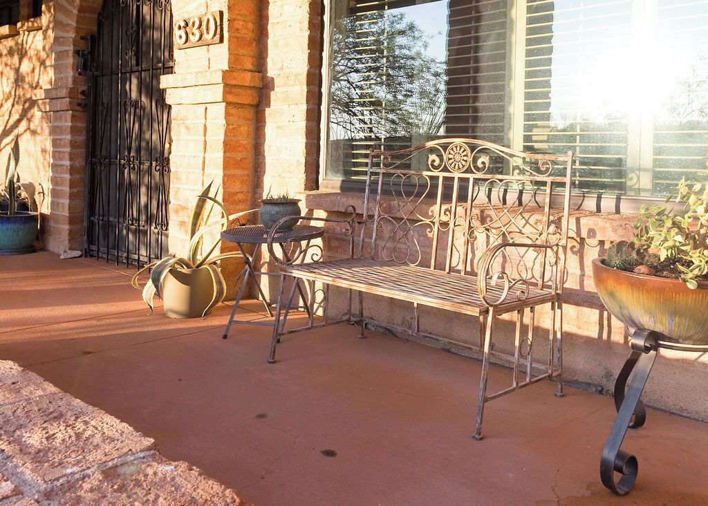 An outdoor seating area with a wrought iron bench and table, potted plants, and a building entrance with a number sign.