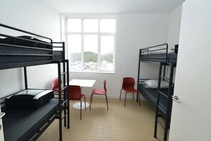 A dormitory room with bunk beds, a small table, two red chairs, and a window with a view of parked cars and greenery. - Schotte Youth Accommodation Centre (Aalst)