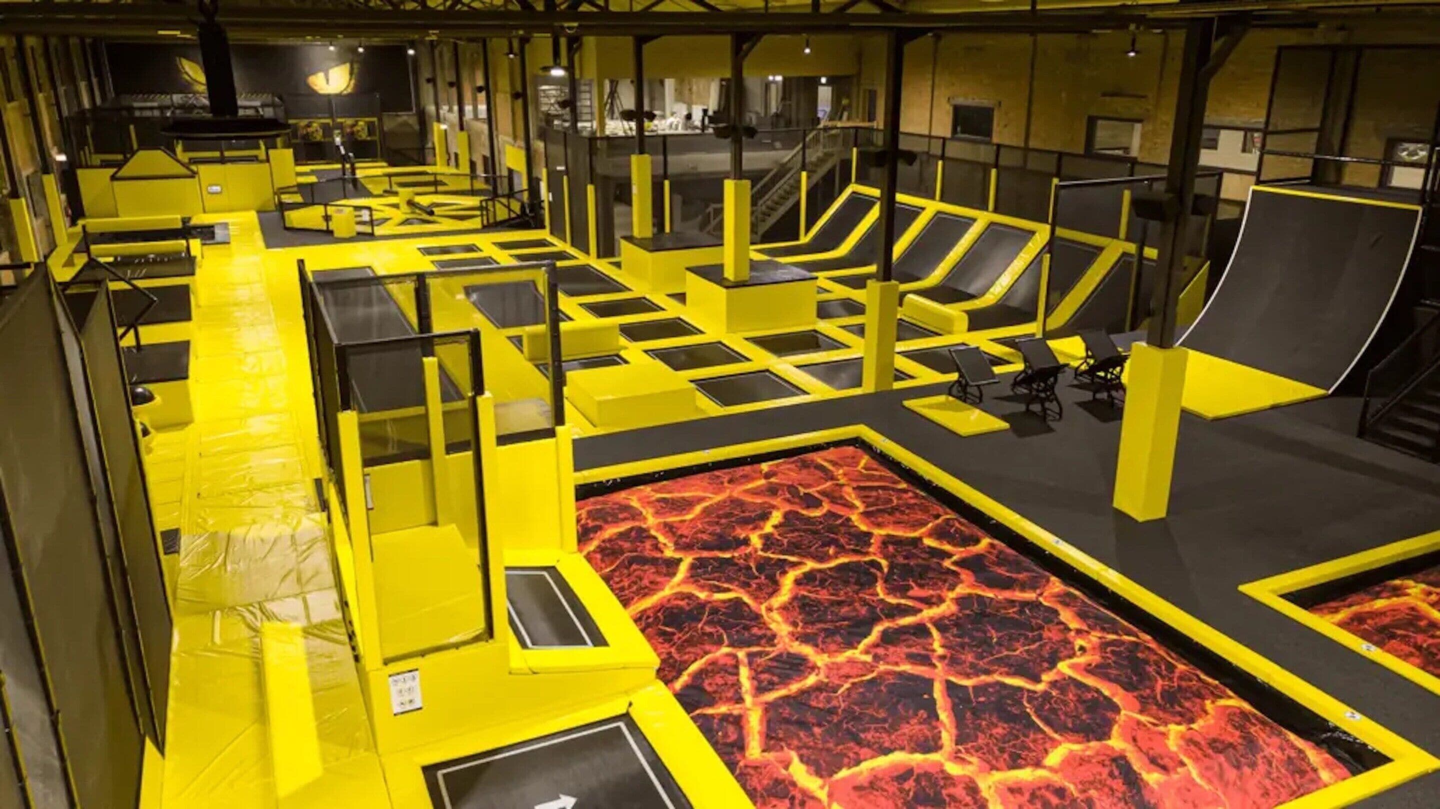 Parque de trampolines interior con barreras de seguridad amarillas y negras, suelo con temática de lava y zonas de descanso.