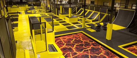 Parque de trampolines interior con barreras de seguridad amarillas y negras, suelo con temática de lava y zonas de descanso.