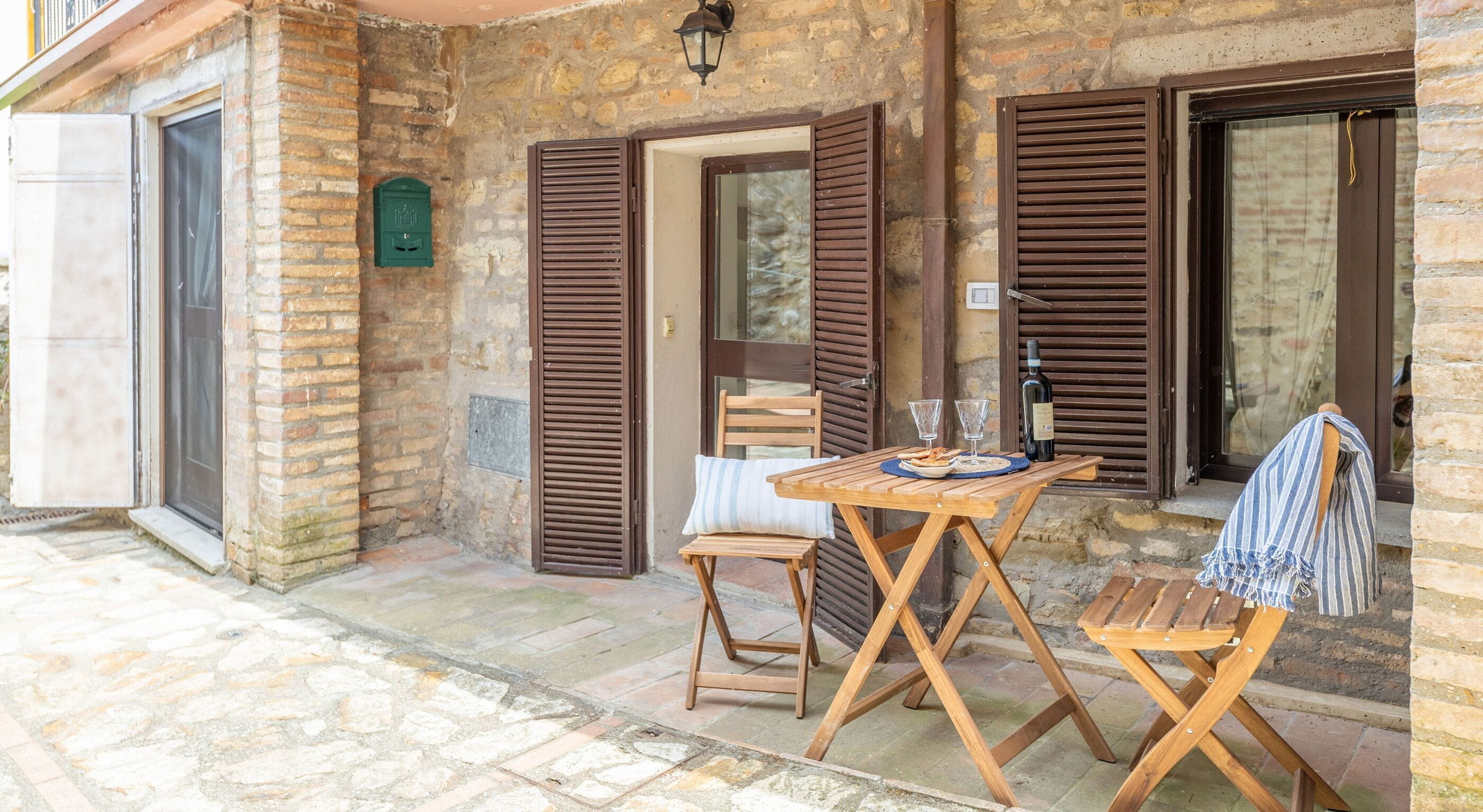 Un bâtiment en pierre avec des volets en bois, une table avec des chaises et une bouteille de vin.
