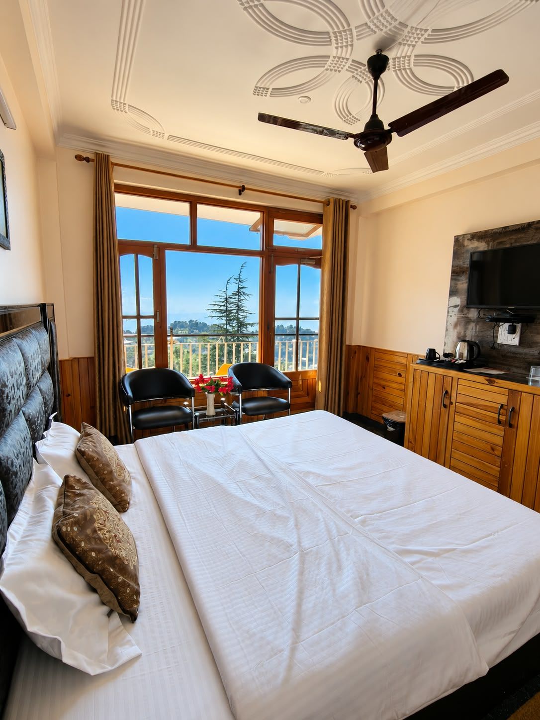 Ein Hotelzimmer mit einem großen Bett, einem Deckenventilator, einem Fernseher und einem Balkon mit Blick auf Bäume.