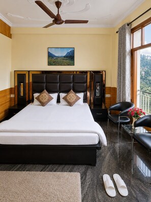 A hotel room with a large bed, a ceiling fan, a painting on the wall, a window with a view, and a pair of slippers on the floor.