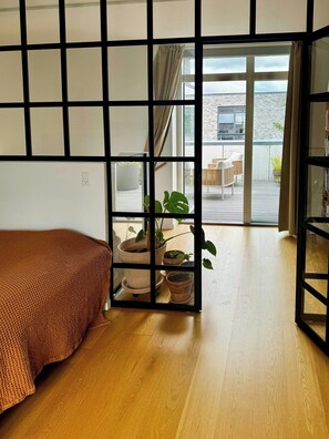 Une pièce avec un plancher en bois, un lit recouvert d’une couverture marron et une porte-fenêtre coulissante donnant sur un espace extérieur agrémenté de plantes et d’un coin salon.