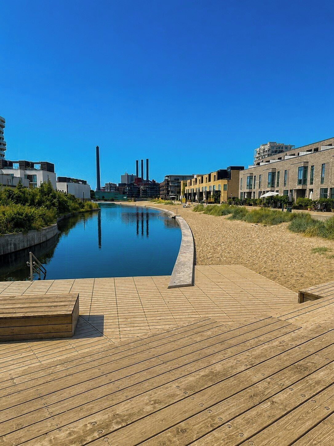 A swimming pool with a wooden deck, surrounded by buildings and a clear sky.