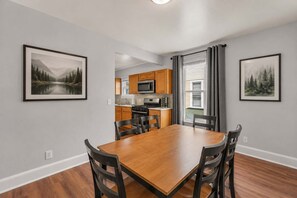 A dining area with a wooden table and chairs, a kitchen with wooden cabinets, and framed landscape paintings on the walls. - Clubhouse - All Star Baseball Rentals (Oneonta)