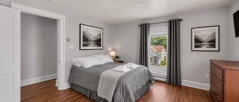 Une chambre à coucher comprenant un lit, une table de chevet, une commode, deux tableaux encadrés, une fenêtre avec des rideaux et une porte.