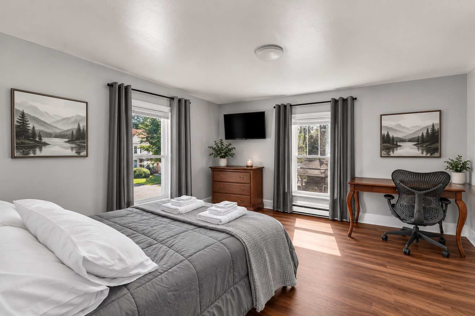 A bedroom with a bed, a desk, a chair, a TV, and two windows with curtains.