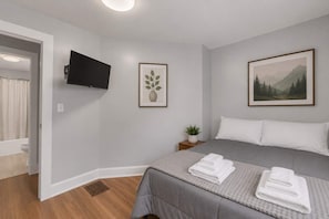 A hotel room with a bed, a television mounted on the wall, a framed picture, and a small bedside table with a plant. - Field House - All Star Baseball Rentals (Oneonta)