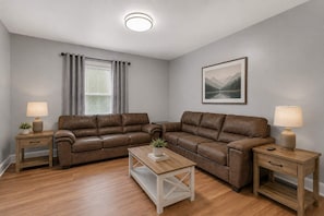 A living room with a brown leather sectional sofa, a coffee table, two nightstands with lamps, a window with curtains, and a framed picture on the wall. - Field House - All Star Baseball Rentals (Oneonta)
