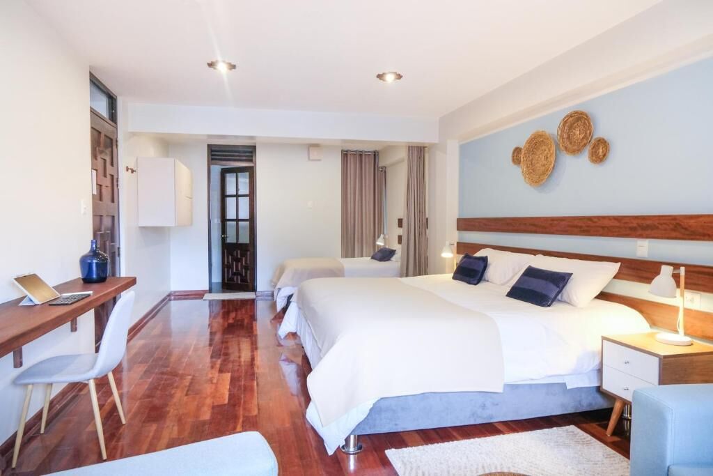 A hotel room with a wooden floor, a desk with a chair, a bed with white bedding, and a nightstand with a lamp.
