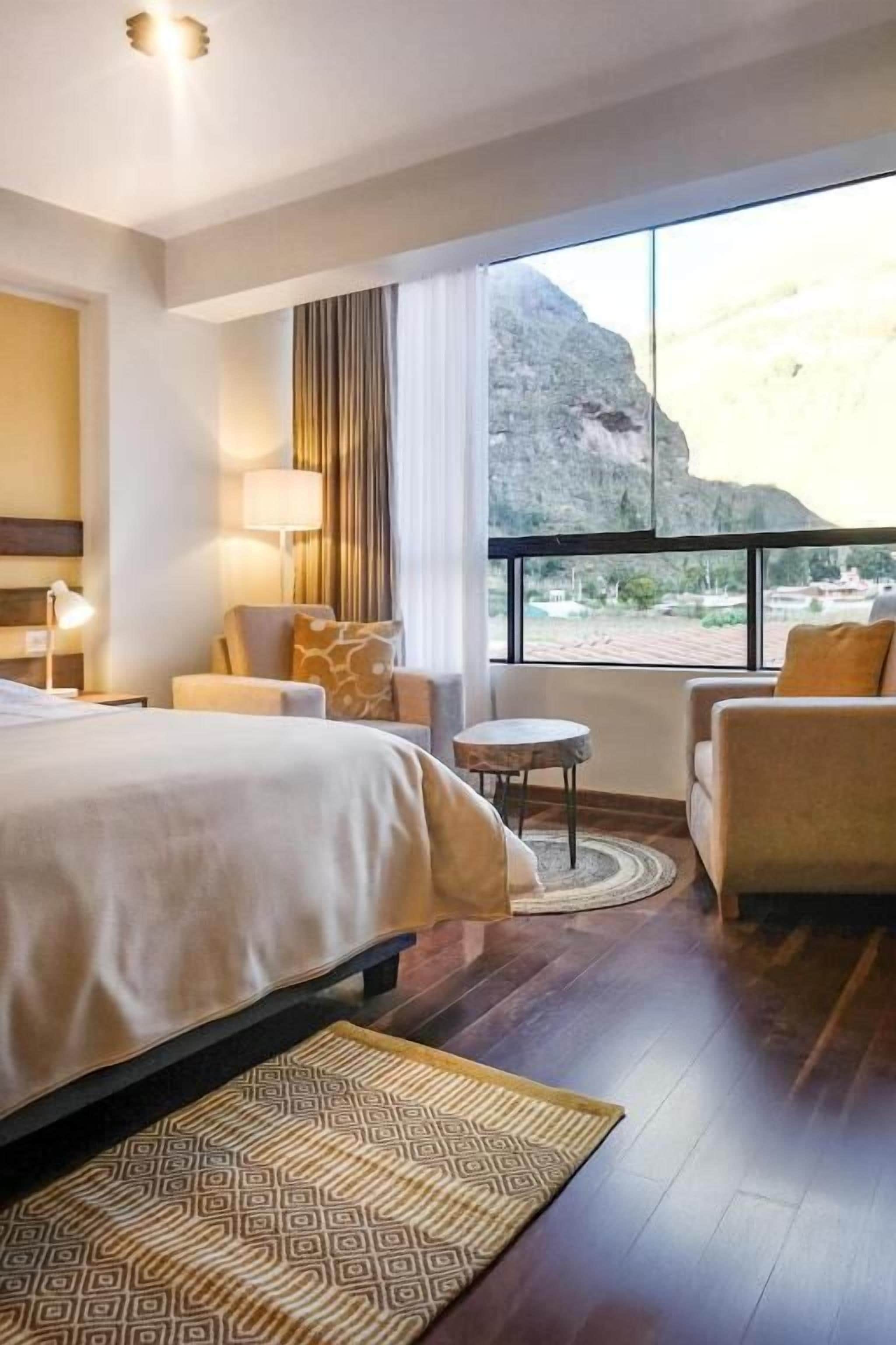 A hotel room with a bed, a small table, a chair, and a view of a mountain.