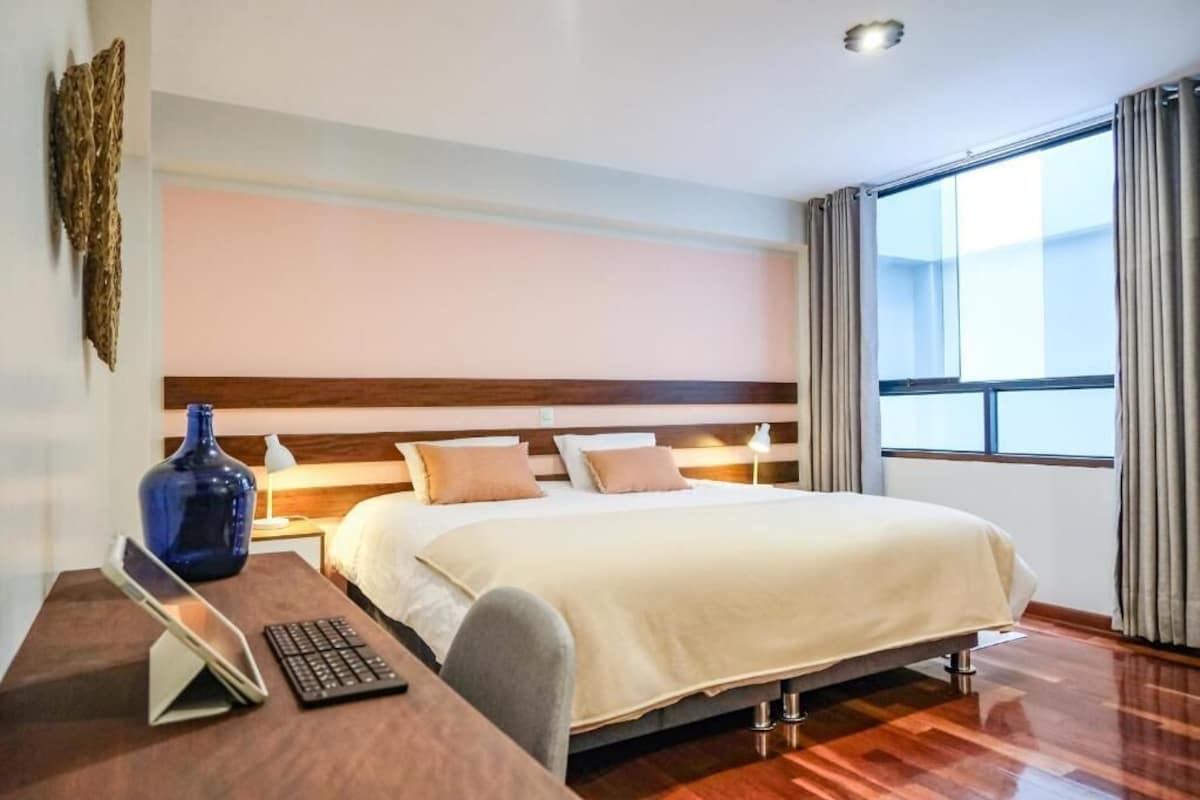 A hotel room with a bed, bedside table, lamp, and a large window with curtains.