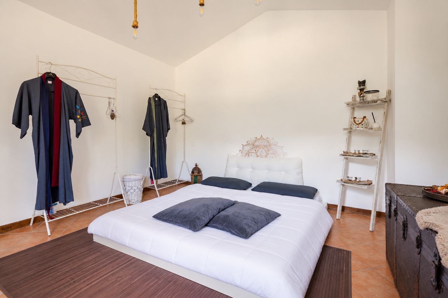 A bedroom with a large bed, a wardrobe, a mirror, a shelf with decorative items, and a suitcase.