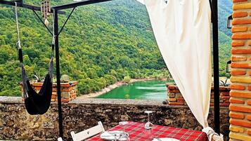 Ein Essbereich im Freien mit einer rot karierten Tischdecke, weißen Stühlen und Blick auf ein grünes Tal.