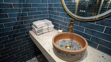 A bathroom with a blue-tiled wall, a round sink with a decorative basin, and a stack of towels on a shelf.