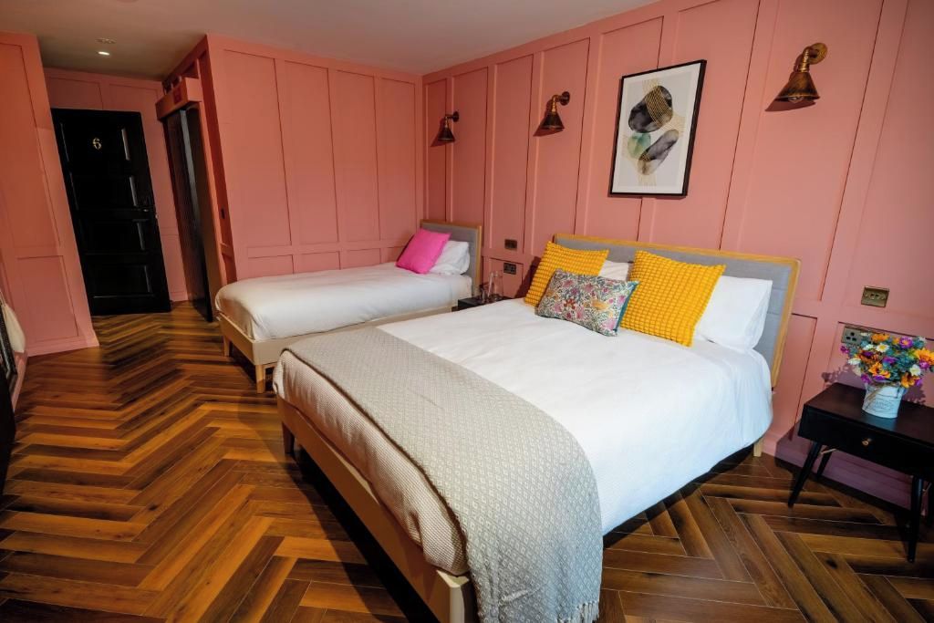 A hotel room with two beds, a wooden herringbone floor, pink walls, and a small bedside table with a vase of flowers.