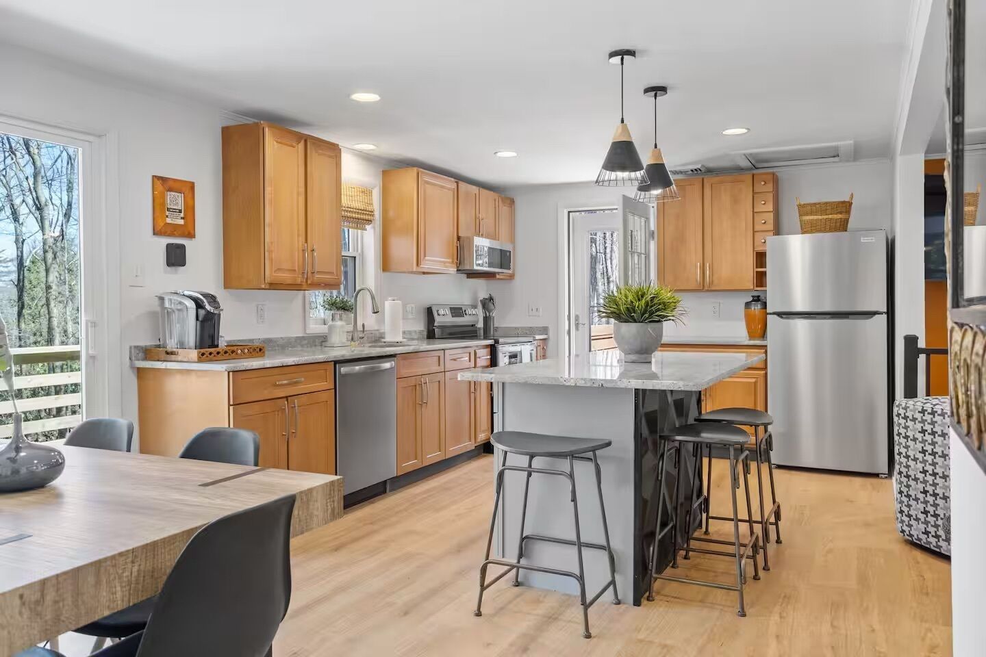 A modern kitchen with wooden cabinets, a central island with stools, stainless steel appliances, and a large window.