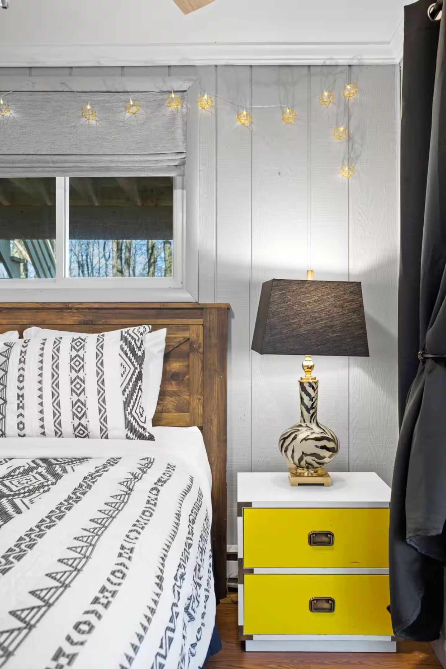 A bedroom with a bed, a nightstand with a lamp, and a window with curtains.