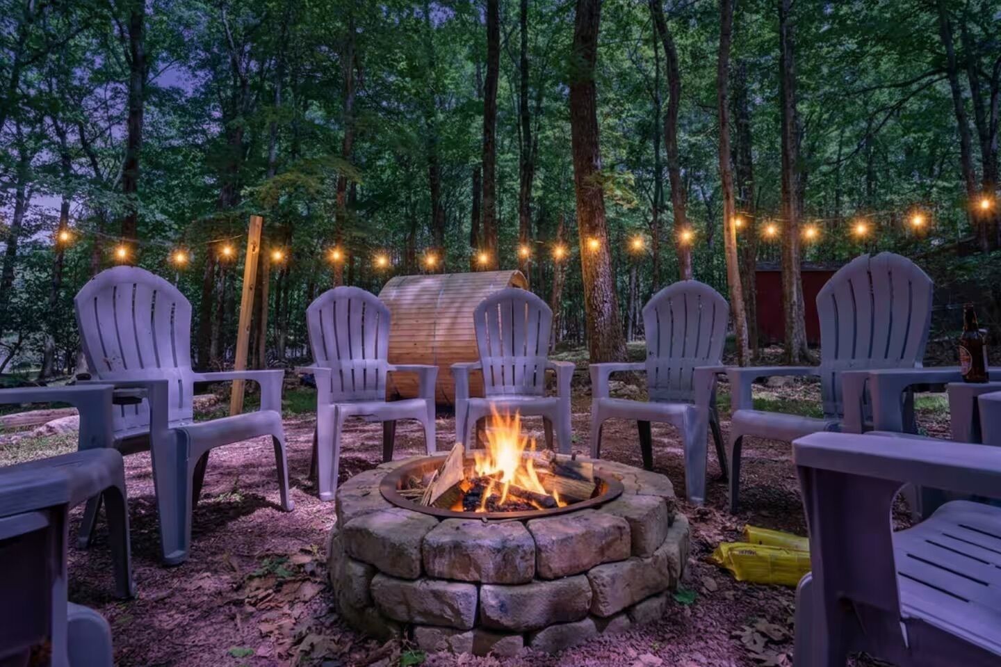 A campfire surrounded by Adirondack chairs in a forest setting.