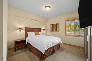 A bedroom with a bed, bedside lamps, a nightstand, a window with blinds, and a wall-mounted artwork. - Yocum Ridge Condo 204 at South Grand Lodge | Best Mt. Hood View (Government Camp)