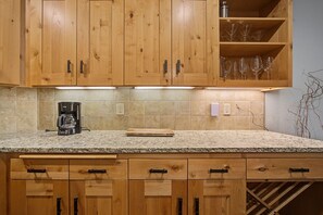 A kitchen with wooden cabinets, a granite countertop, a coffee maker, and wine glasses on a shelf. - Yocum Ridge Condo 204 at South Grand Lodge | Best Mt. Hood View (Government Camp)