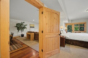 Un dormitorio con puerta de madera, una cama, una mesita de noche, una lámpara y una ventana con vistas a árboles.
