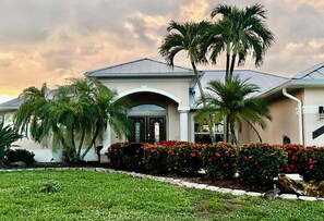 A residential building with a symmetrical facade, a central entrance with an arch, and a well-maintained garden with palm trees and flowering plants. - Hidden Gem in Cape Coral with heated pool and spa, on wide canal 3b/2b (Cape Coral)