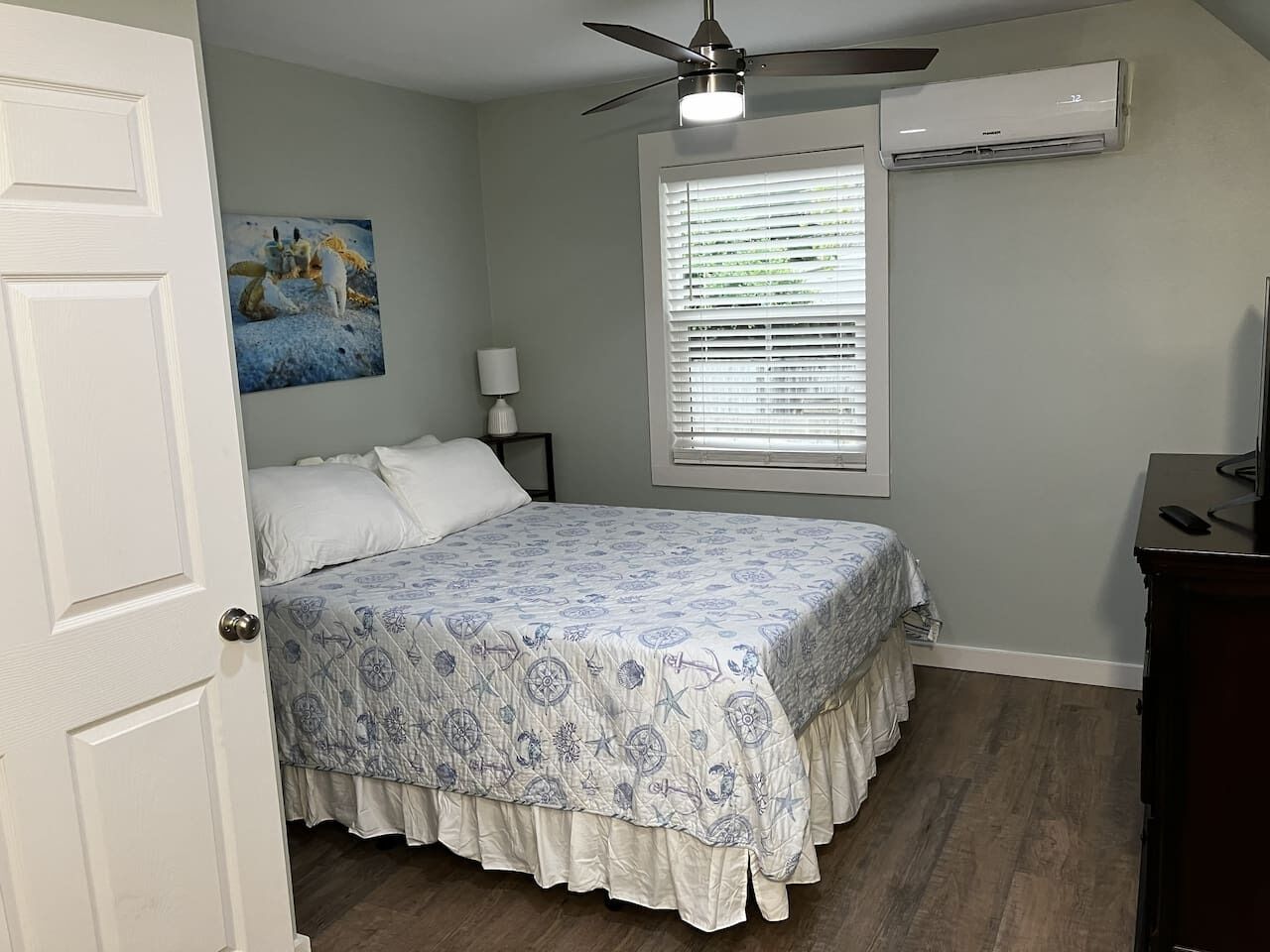 A bedroom with a bed, a ceiling fan, an air conditioning unit, a window with blinds, and a painting on the wall.
